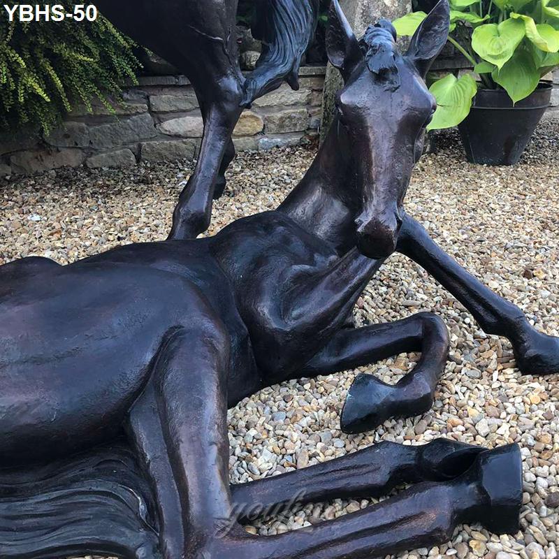 Bronze sculpture of a reclining foal displayed on a gravel garden with lush plants and a stone wall backdrop.