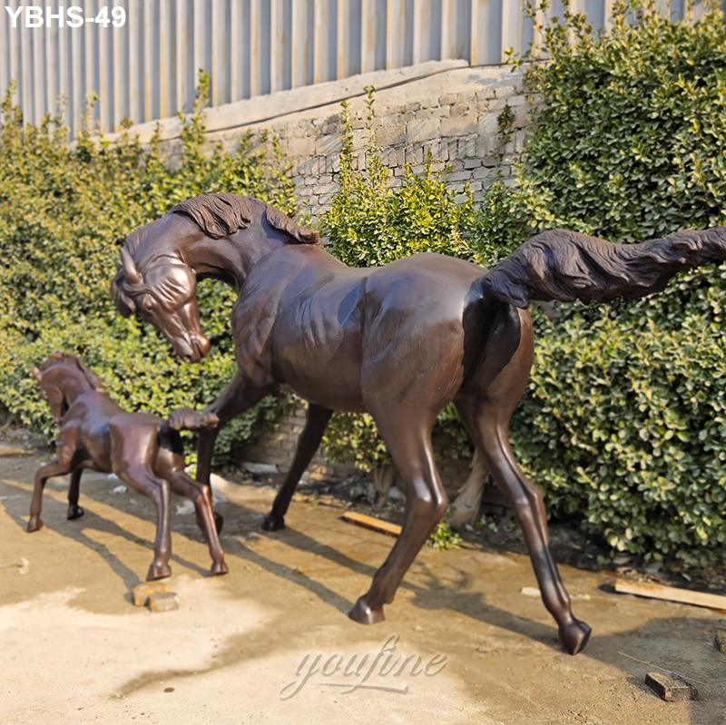 Large bronze horse statue interacting with a smaller foal sculpture, set outdoors near a greenery-lined stone wall.