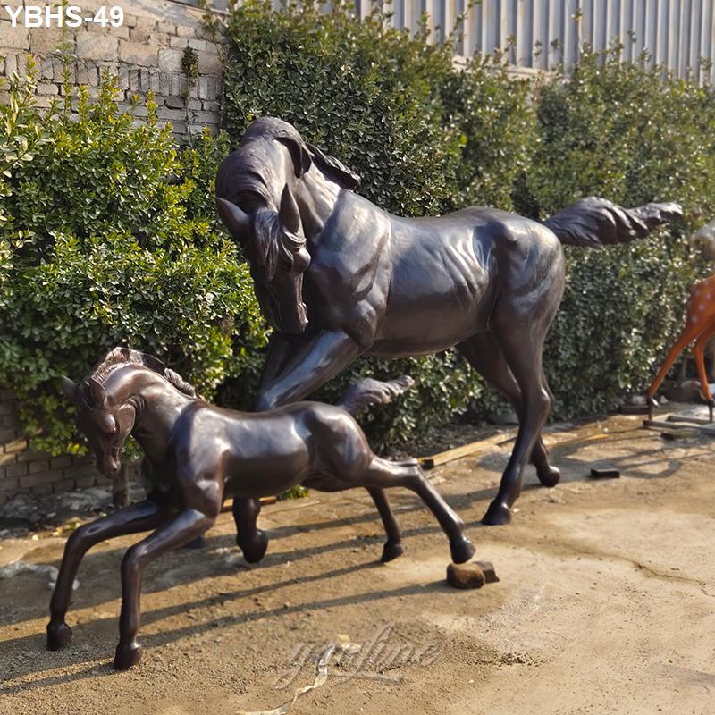 A lifelike bronze sculpture of a horse and foal outdoors, set against greenery and a stone wall. The adult horse appears protective.