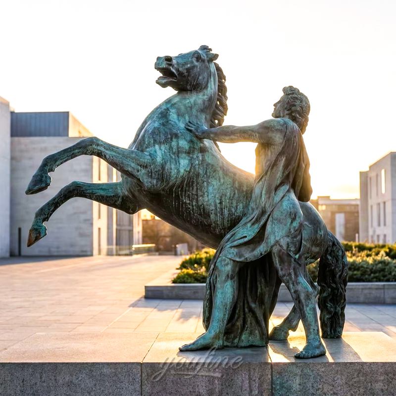 A bronze sculpture of a rearing horse and a rider standing beside it, illuminated by the soft light of sunrise in an urban outdoor setting.