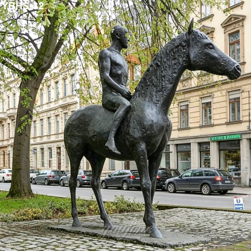 Bronze statue depicting a person sitting on a horse, placed in an urban park surrounded by buildings and cars.