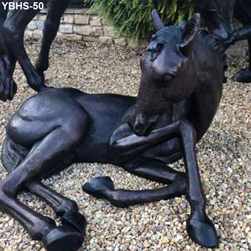 Detailed bronze sculpture of a foal lying reclined on gravel, with greenery and a stone wall in the background.