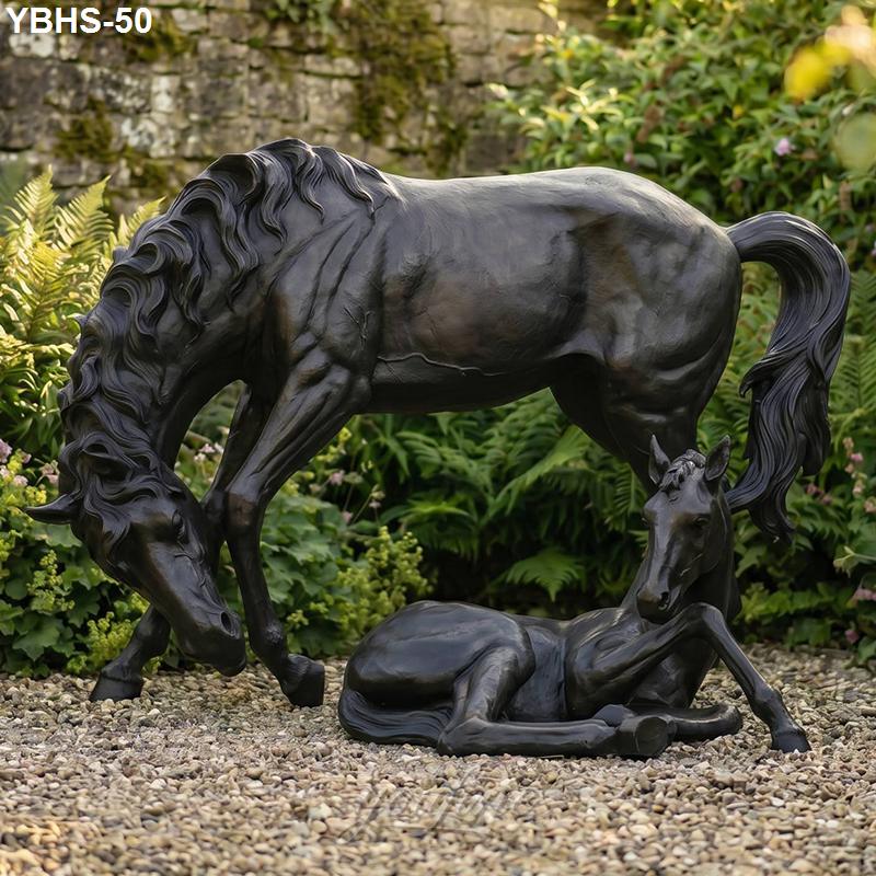 Detailed bronze statue of a horse standing and its foal resting on gravel, surrounded by lush greenery in a garden setting.