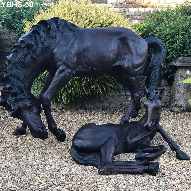 Elegant bronze horse sculpture featuring a mare grazing and a foal resting on gravel, surrounded by shrubs and a rustic stone wall.
