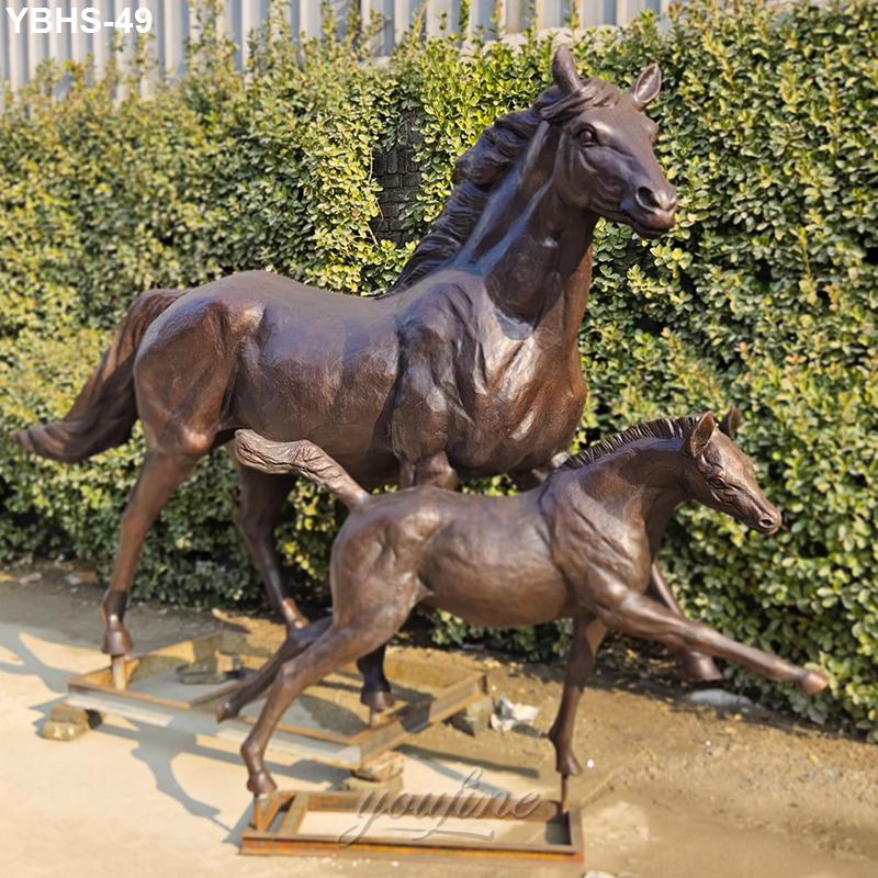 Bronze sculptures of a mare and foal in dynamic poses, set outdoors on a concrete base against a green hedge background.