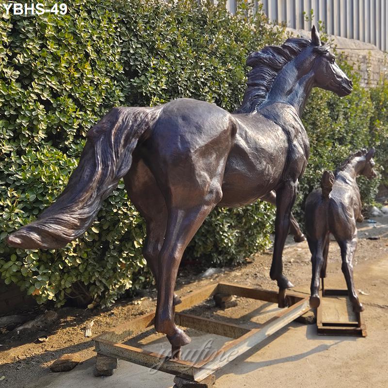A pair of bronze horse sculptures, an adult horse and a foal, displayed outdoors on stands with greenery in the background.