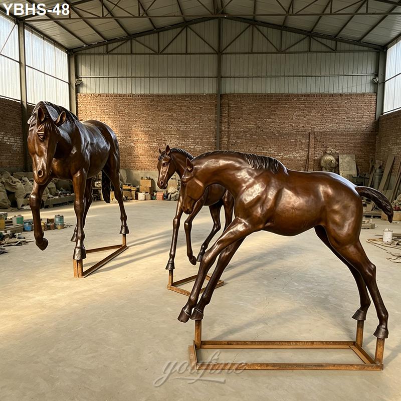 Three bronze horse sculptures on support frames inside a spacious workshop with brick walls and high ceilings.