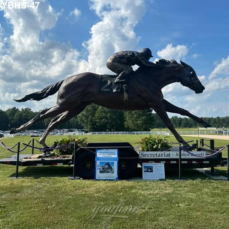 Bronze statue of Secretariat depicted racing with a jockey, displayed outdoors on a trailer surrounded by informational boards.