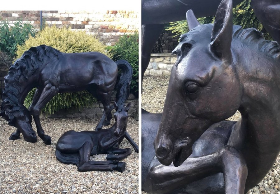 Elegant bronze sculpture of a horse bending over its lying foal, displayed on gravel in a landscaped garden with shrubbery.