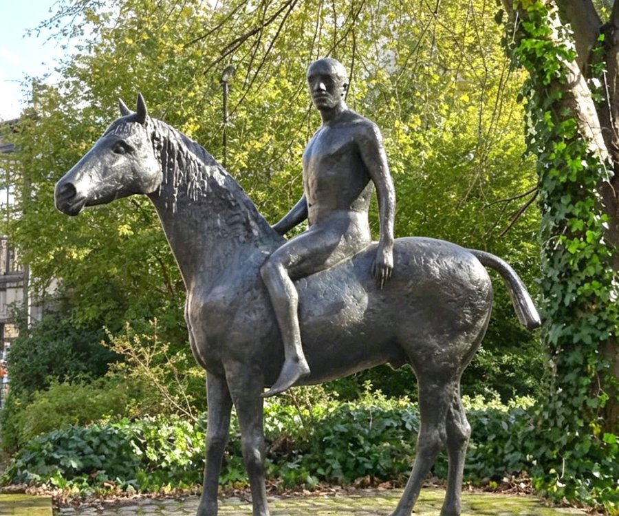 A bronze statue of a rider on horseback in a lush park setting with trees and greenery in the background.