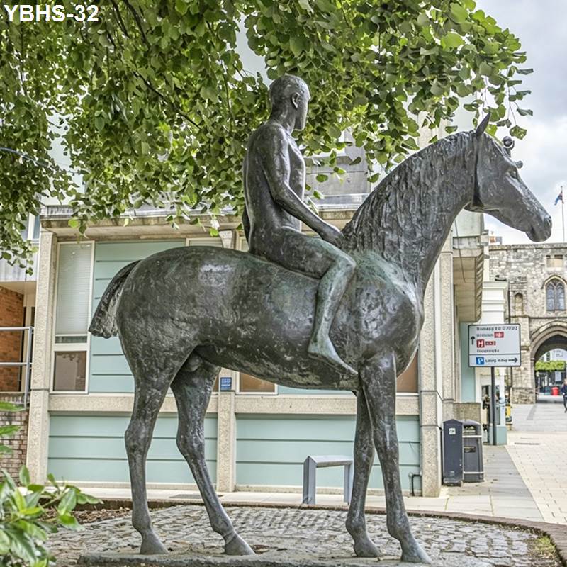 Bronze statue of a seated male rider on a horse under a leafy tree, with urban buildings and cobblestone ground in the background.