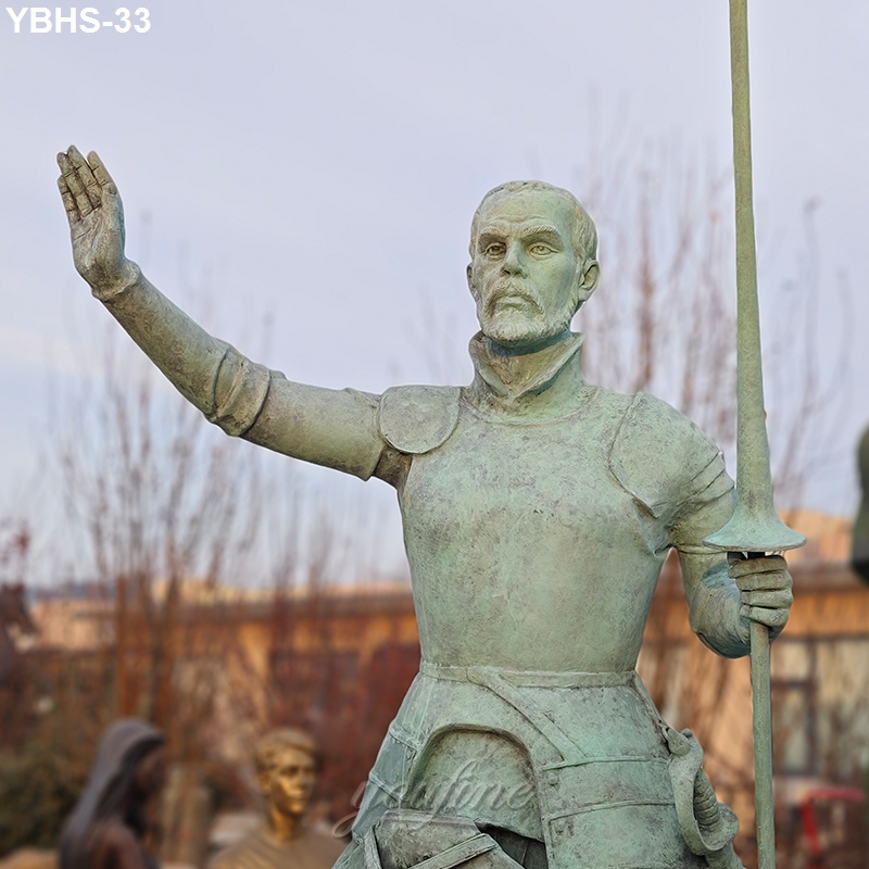 A bronze statue of a knight in armor holding a spear in one hand while raising the other arm, set against an outdoor backdrop.
