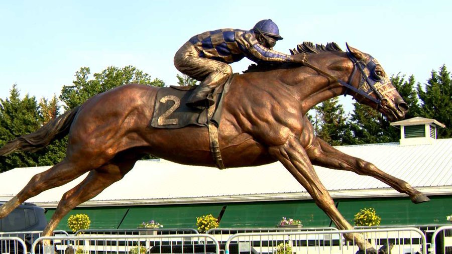 Detailed bronze statue of a racehorse in motion with jockey, displayed outdoors near a building and greenery.