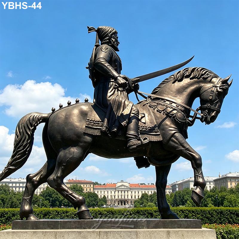 A detailed bronze statue of a warrior riding a horse, holding a curved sword, with a vibrant blue sky and historic buildings in the background.