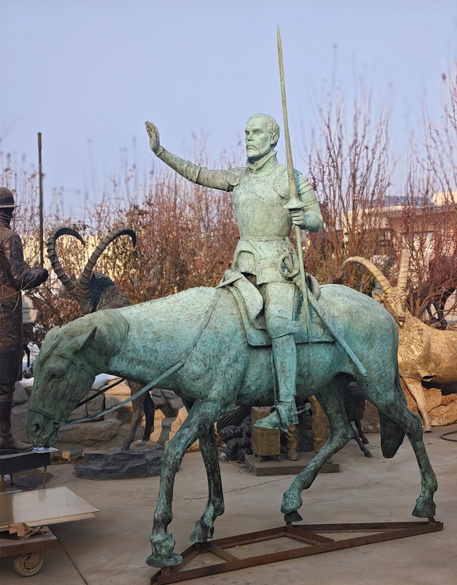 Bronze statue of a warrior on horseback holding up a sword, with detailed armor and reins. Background shows trees and other figurines.