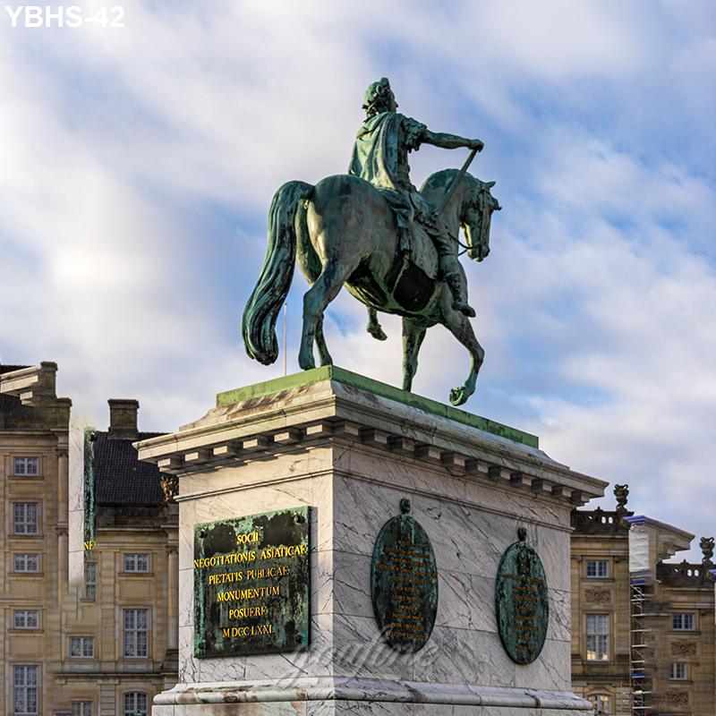 Equestrian bronze statue depicting a rider on horseback atop a marble pedestal with engraved plaques, set against historic buildings and a blue sky.