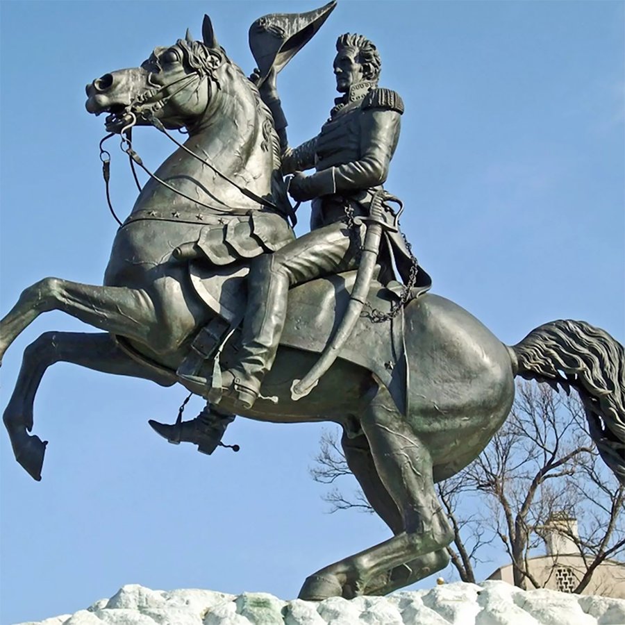 Bronze equestrian statue showing a rider in military attire holding a hat, with the horse rearing on its hind legs, set against a clear blue sky.