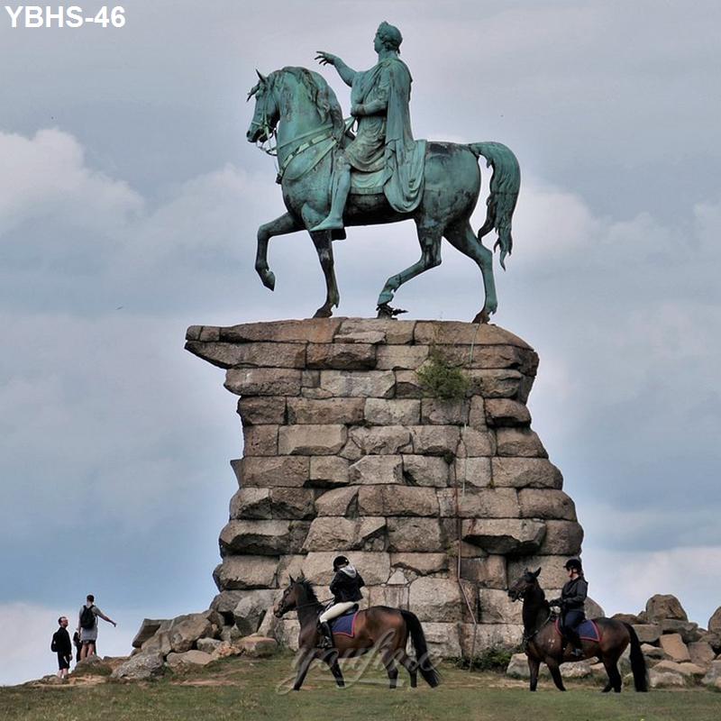 Bronze equestrian statue on a tall stone pedestal with two horseback riders in the foreground and visitors on foot nearby.