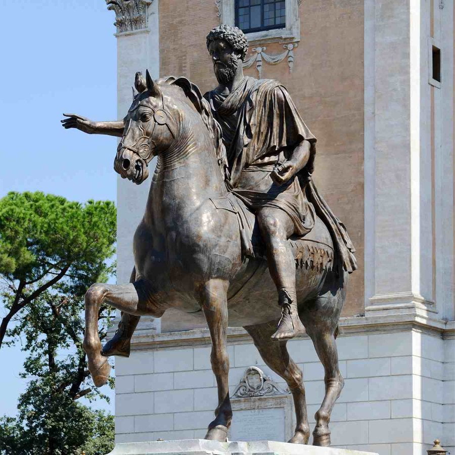 Bronze Marcus Aurelius statue of a historical figure with an outstretched arm, positioned in front of an ornate building and trees.