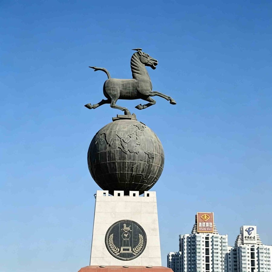 Bronze Flying Horse of Gansu is poised atop a globe-shaped pedestal with detailed map engravings, set against a clear blue sky with buildings in the background.
