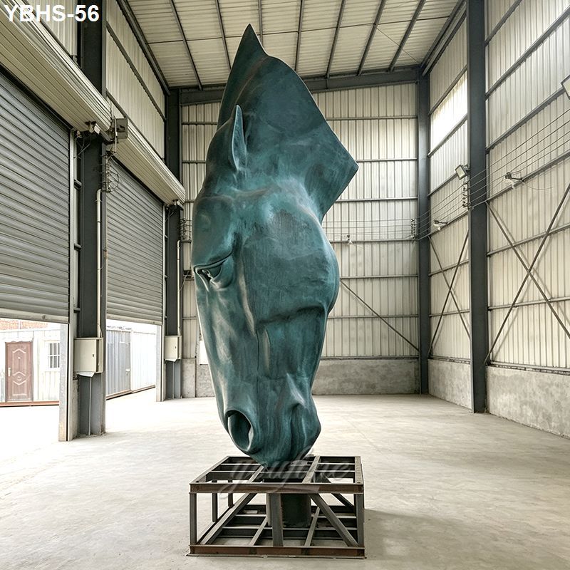 A large, patinated bronze horse head sculpture placed on a metal base inside a spacious industrial warehouse.