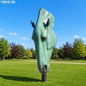 Large green patina bronze horse head sculpture set on a manicured lawn in a park with clear blue skies and trees in the background.
