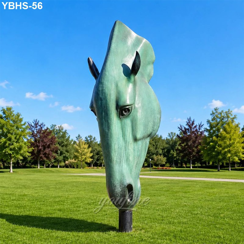 Large green patina bronze horse head sculpture set on a manicured lawn in a park with clear blue skies and trees in the background.