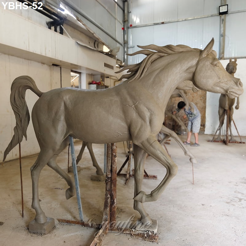 Clay mold of a detailed horse sculpture supported by frames, in a workshop setting with an artisan working in the background.