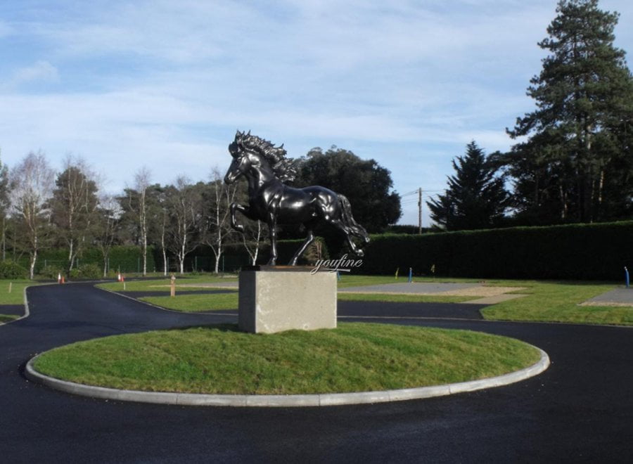 A shiny black bronze horse sculpture stands on a raised stone pedestal in the center of a landscaped roundabout with paved roads and green grass.
