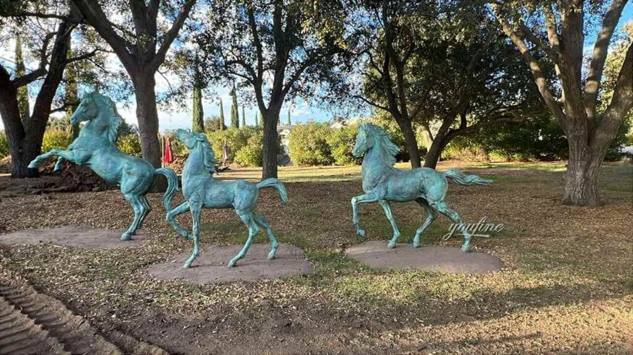 Three bronze horse sculptures with galloping poses placed on a grassy area surrounded by trees, creating a serene outdoor setting.