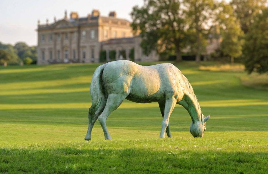 Bronze horse sculpture grazing on a lush green lawn with a grand manor in the background surrounded by trees.