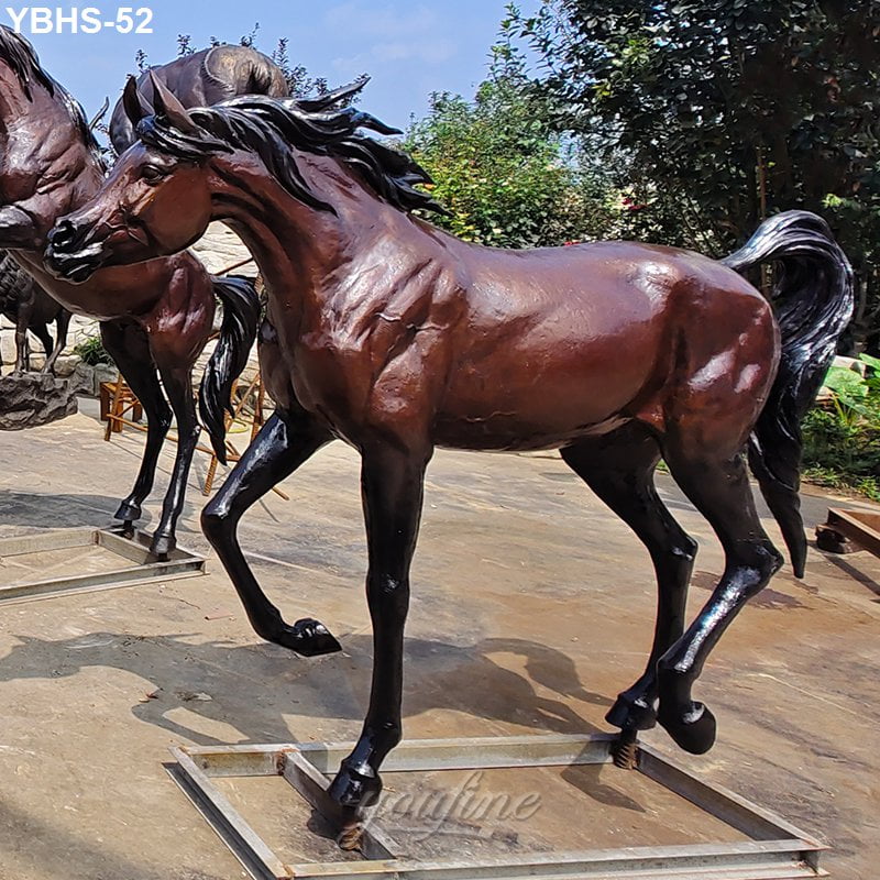 A detailed bronze Arabian horse statue in a dynamic pose, set outdoors on a paved surface with greenery in the background.