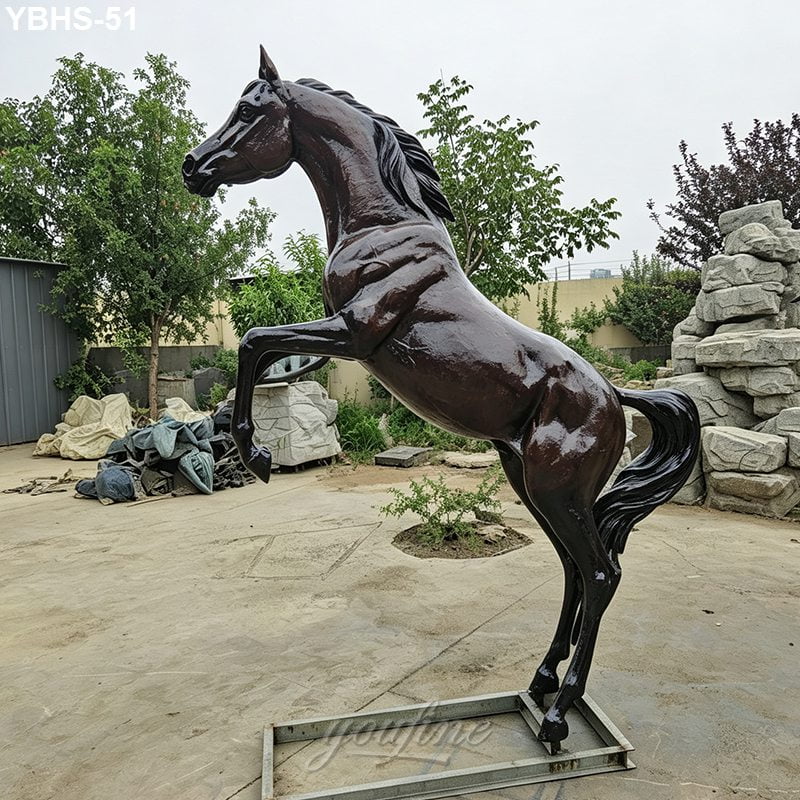 A polished bronze rearing horse sculpture displayed outdoors, surrounded by greenery, stone decor, and industrial elements.