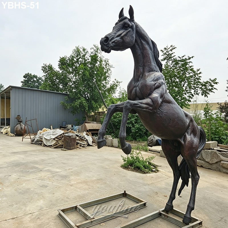 A detailed bronze horse sculpture rearing on its hind legs, displayed in an outdoor setting with trees and a metal shed in the background.