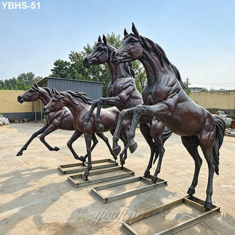 Bronze horse statues in rearing poses placed outdoors on metal mountings, with a backdrop of a fence and greenery under a blue sky.