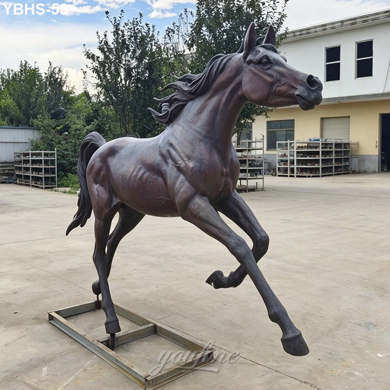 A dynamic bronze Arabian sculpture of a running horse with a muscular build, captured outdoors in a workshop area with trees and buildings in the background.
