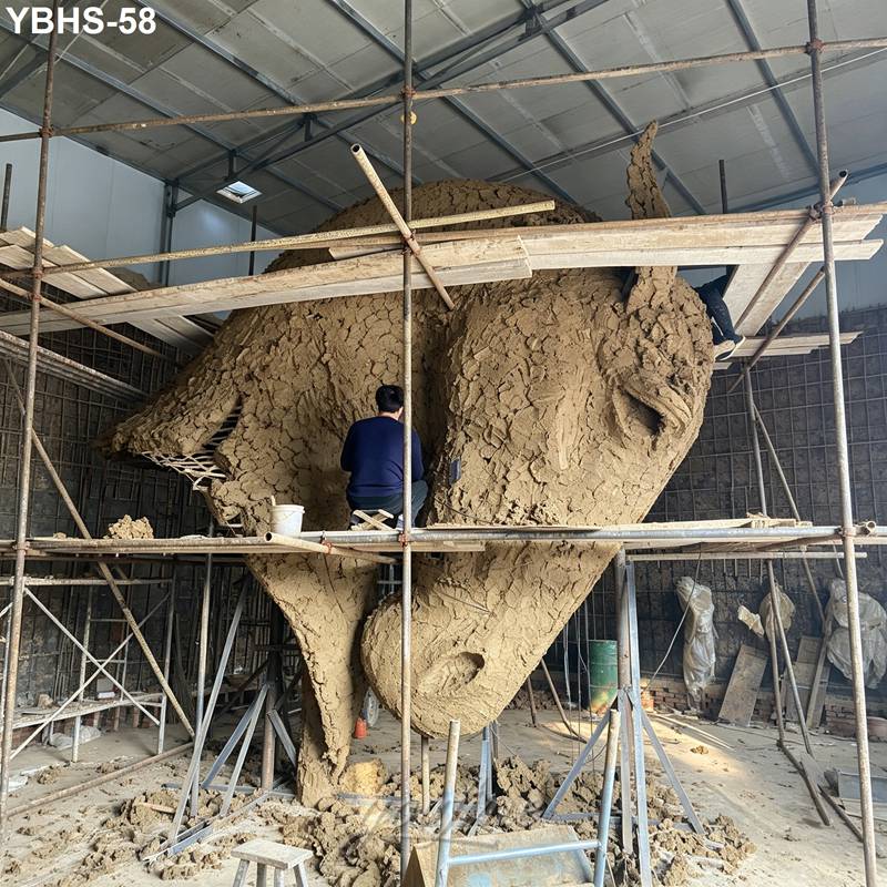 An artist works on shaping a large clay horse head sculpture under scaffolding inside an indoor studio, surrounded by tools and materials.
