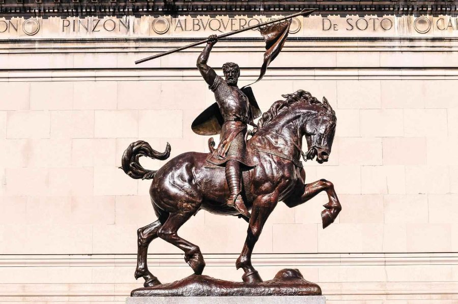 A bronze El Cid Campeador statue depicting a knight holding a flag aloft, mounted on a rearing horse, set against a stone wall.