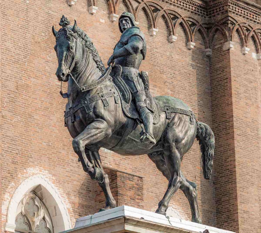 A bronze equestrian statue of Bartolomeo Colleoni featuring a knight in armor on a rearing horse, set against a decorative brick building facade.