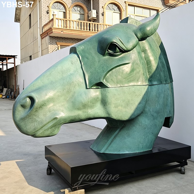A large green patinated bronze horse head sculpture on a black base, situated outdoors near a decorative building.