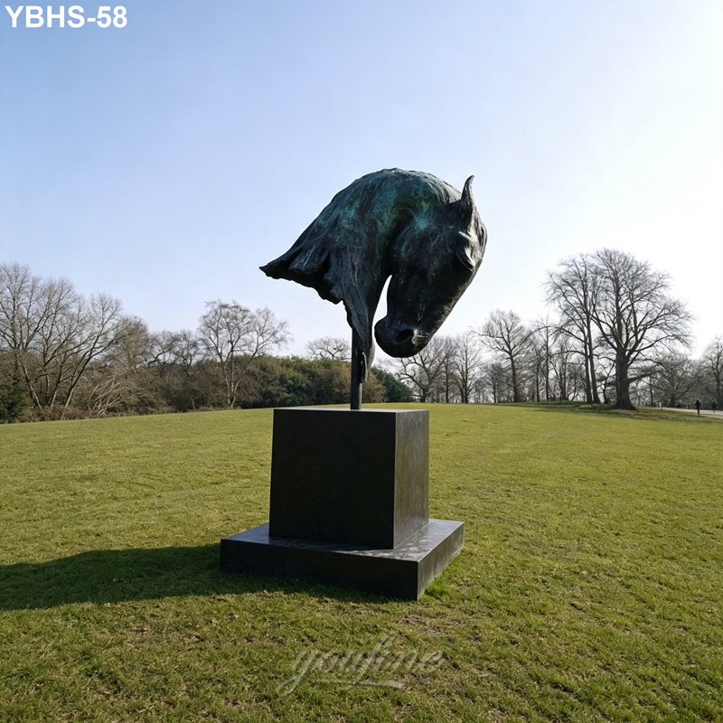 A bronze horse head sculpture on a square pedestal in a grassy open field with scattered trees under a clear sky.