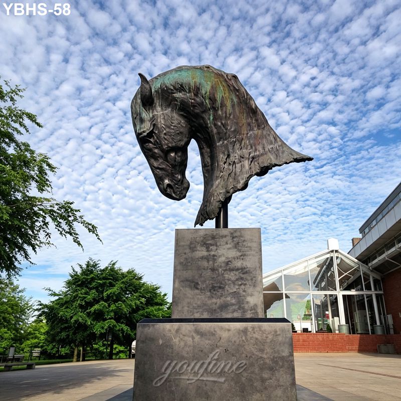 Large bronze horse head sculpture on a stone pedestal, displayed outdoors against a vibrant cloudy sky, with trees and modern architecture nearby.