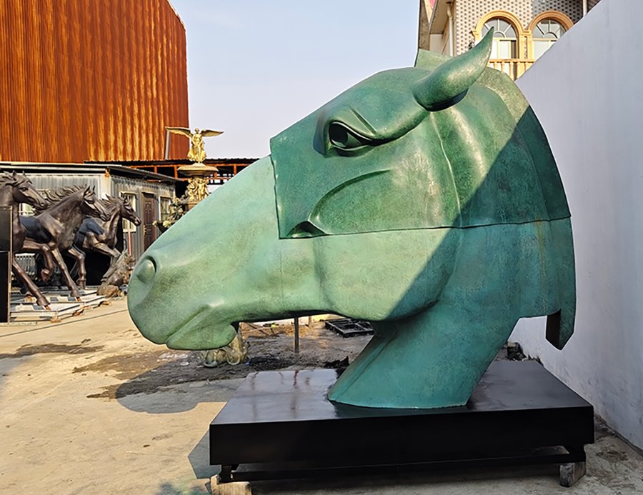 Large green bronze horse head sculpture mounted on a black base in an outdoor area, surrounded by other artistic elements.
