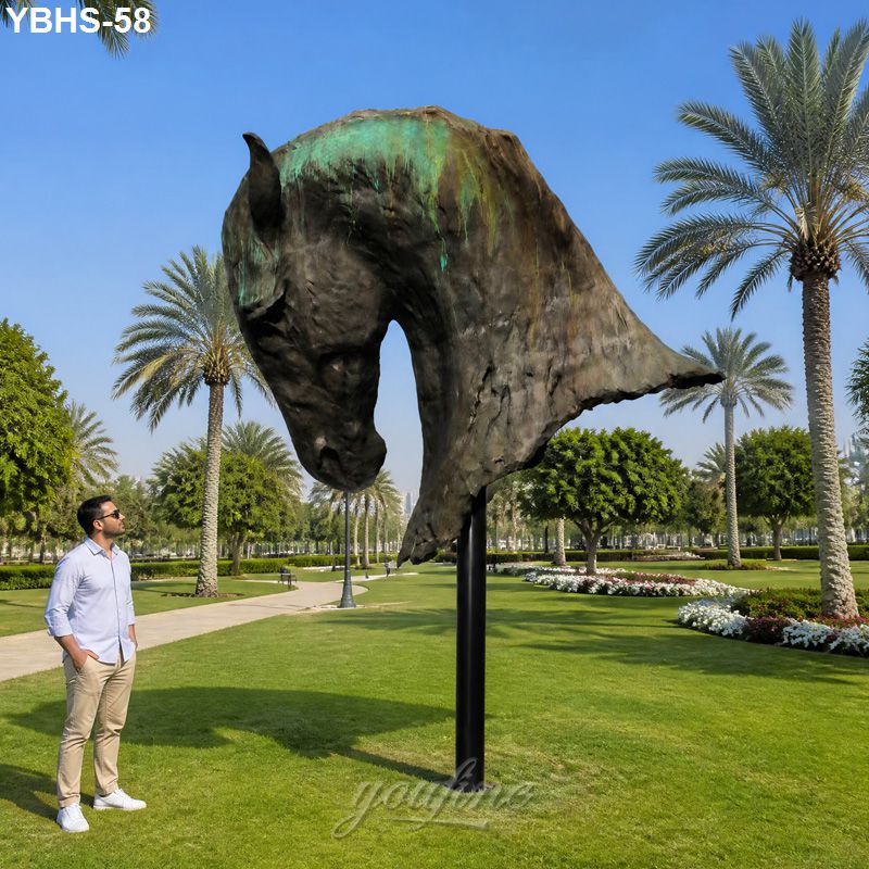 Large bronze horse head sculpture mounted on a pedestal in a landscaped park with palm trees, a man observing it closely.