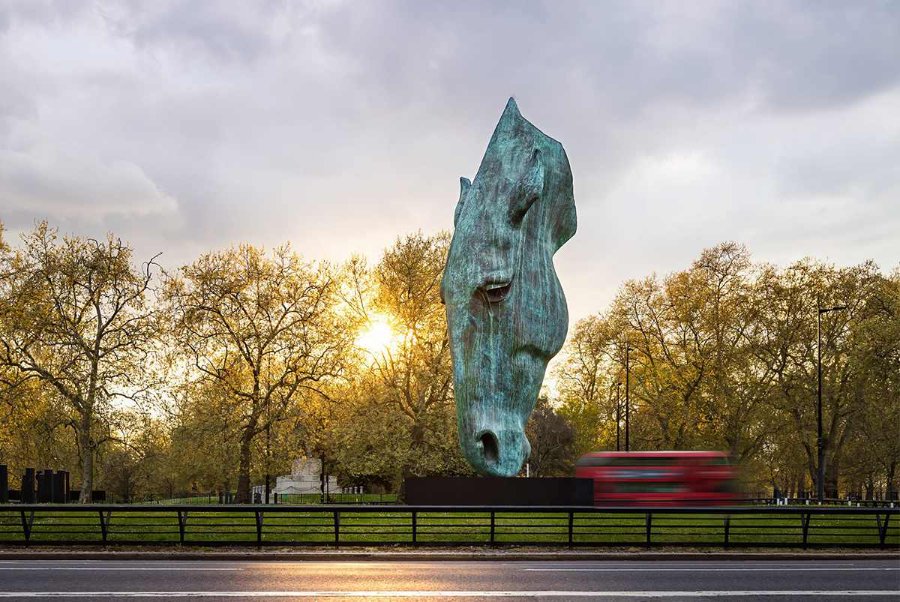 A large bronze horse head sculpture in a park with trees, a setting sun, and a blurred red double-decker bus passing by.