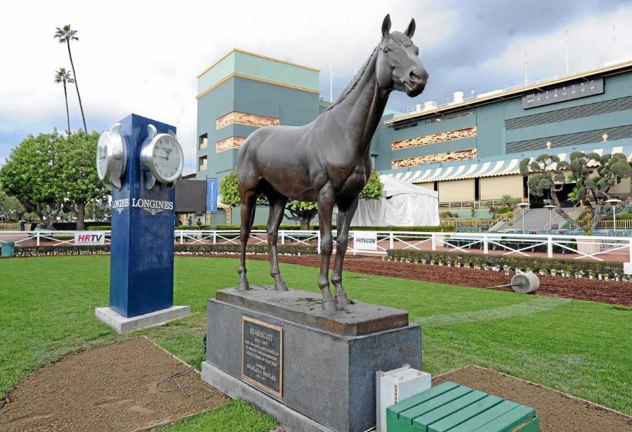 Bronze Seabiscuit horse statue on a pedestal with a commemorative plaque at a racecourse, next to large Longines clock and greenery.