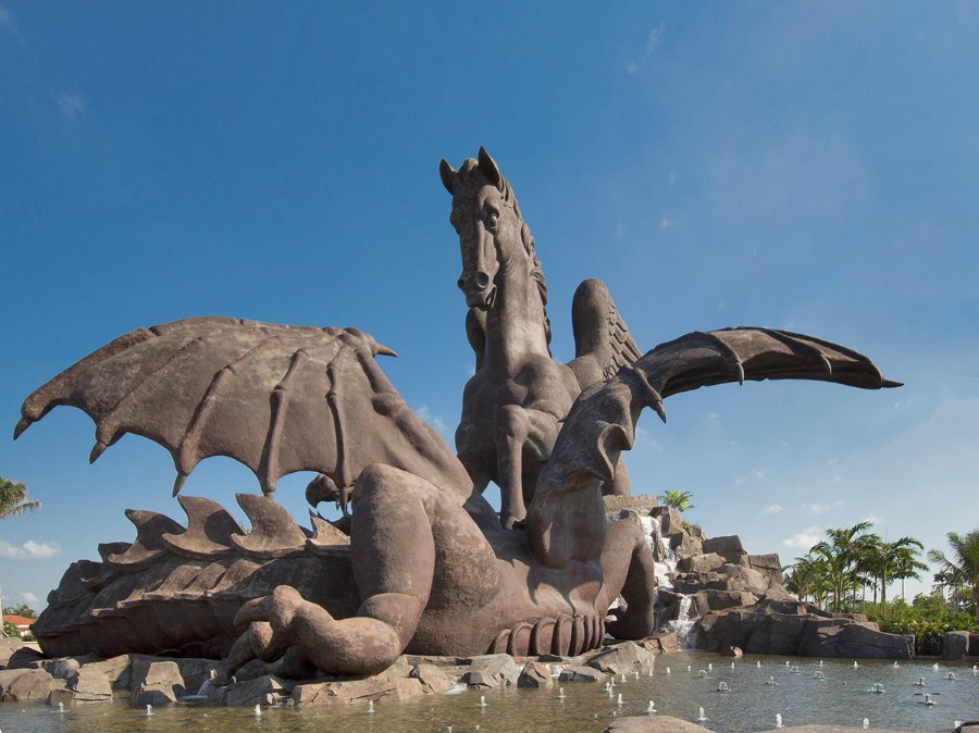 Massive bronze sculpture of Pegasus battling a dragon with wings spread, set in an outdoor landscape with blue skies and water fountain.
