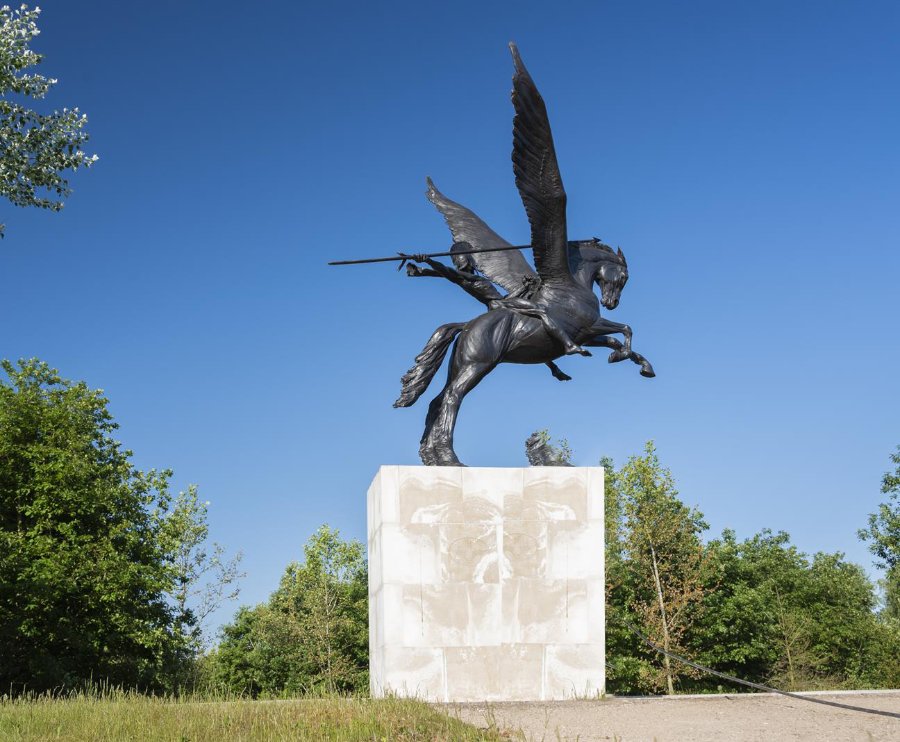Bronze statue of a winged horse with a rider holding a spear, mounted on a stone pedestal against a clear blue sky with surrounding greenery.