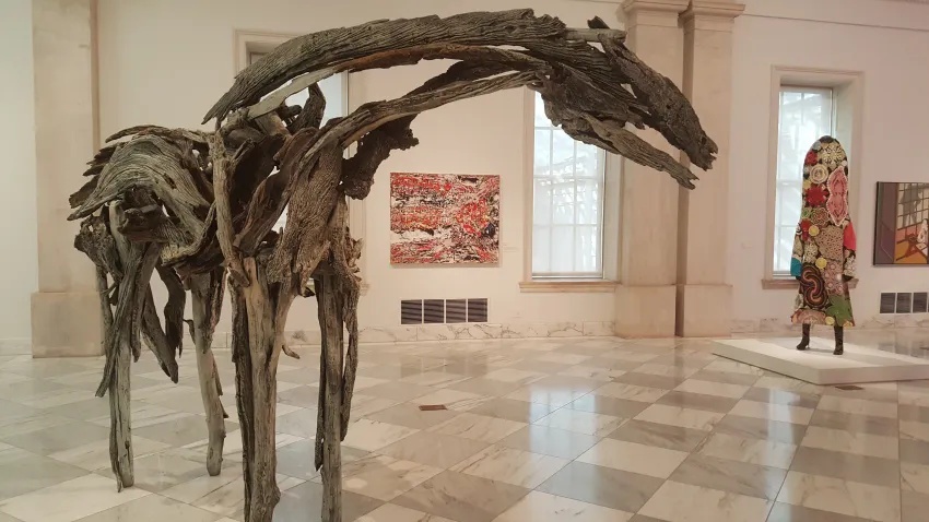A Monekana horse sculpture crafted from driftwood displayed in a marble-floored art gallery with other artworks and a statue in the background.