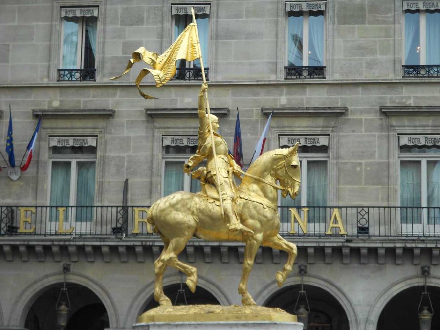 A gilded equestrian Jeanne d'Arc statue holding a raised flag, positioned in front of Hotel Regina with visible balconies and flags.
